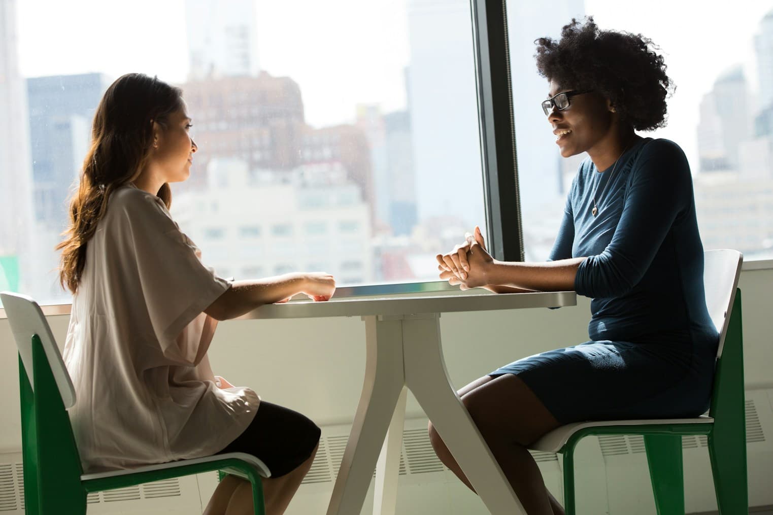 Two people in conversation across a table beside a city window